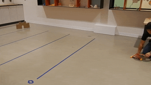 A cork is fired from a K’NEX catapult towards a cardboard target. The cork hits the floor near the target, and bounces past it.   
