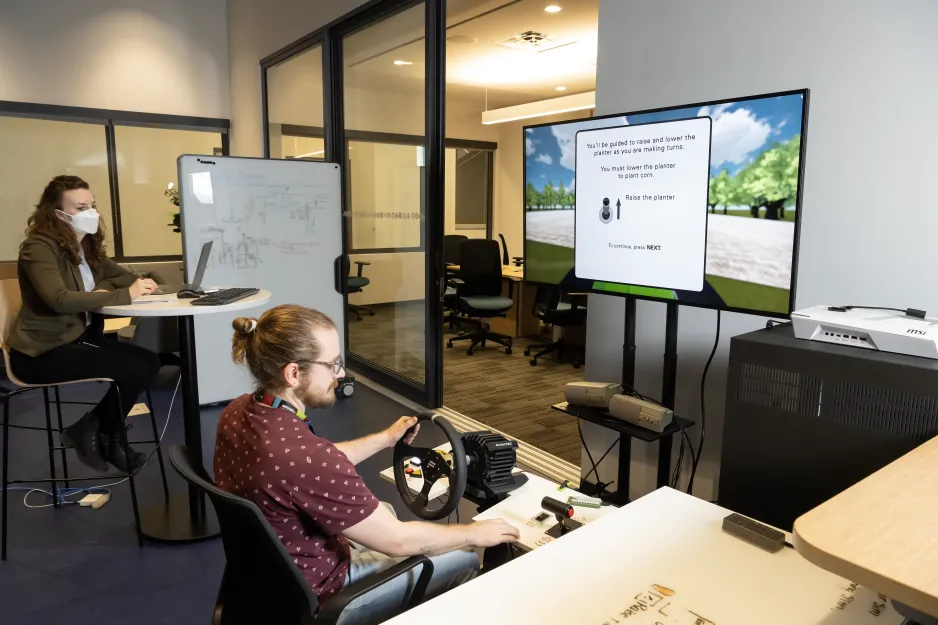 Une personne, les mains sur le volant, regarde une simulation de jeu vidéo avec une deuxième personne qui prend des notes sur un ordinateur portable. En arrière-plan, un tableau blanc avec des notes.