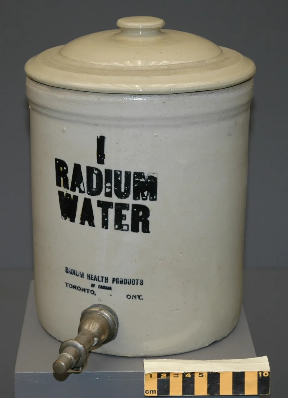 An off-white ceramic jar with ceramic lid and metal spigot at the bottom. The jar has “I RADIUM WATER” and the manufacturer’s information “RADIUM HEALTH PRODUCTS/ TORONTO, ONT.” stenciled on the side in black paint.