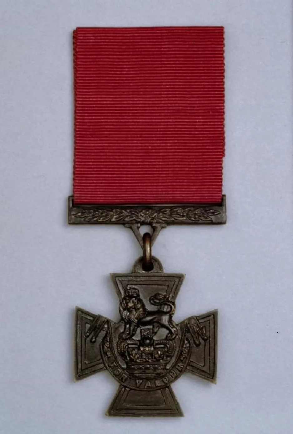 A Victoria Cross medal — a bronze cross with a lion atop a crown, with the inscription “For Valour” across the bottom. 