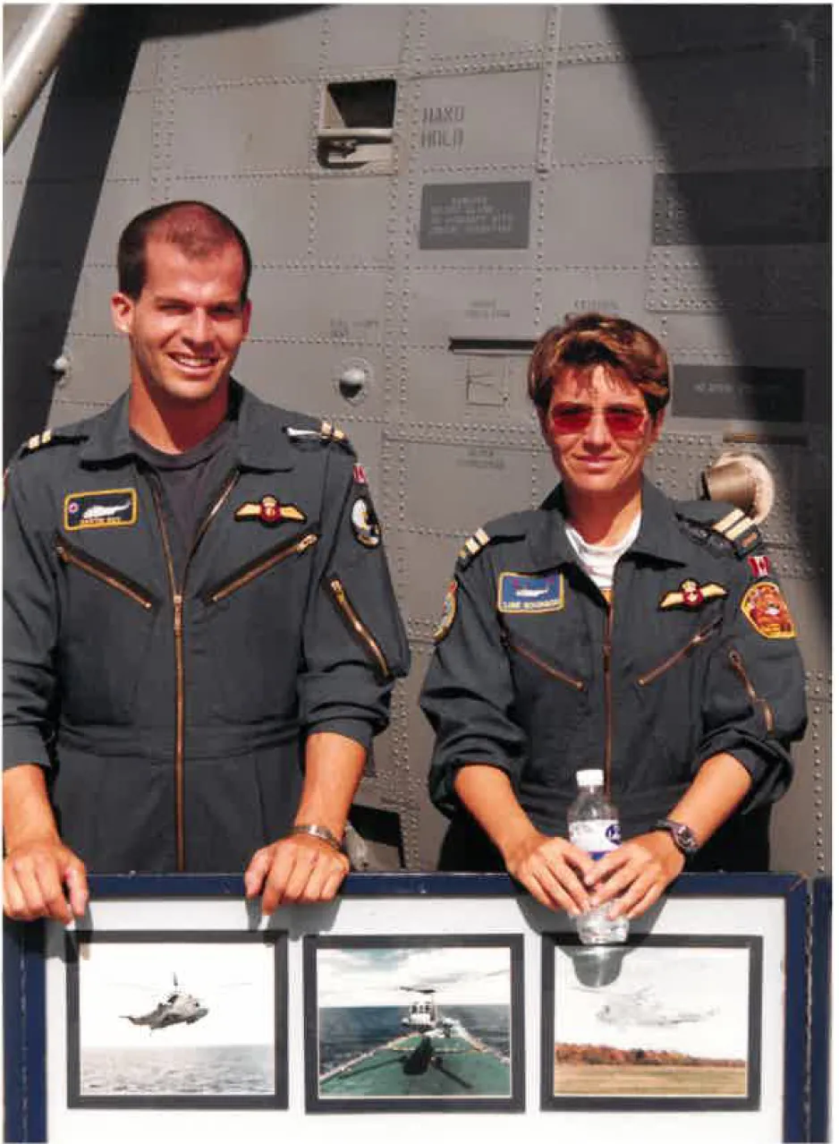 Une femme et un homme en uniforme militaire bleu se tiennent debout derrière un grand cadre comprenant plusieurs photos; on devine un aéronef en arrière-plan.