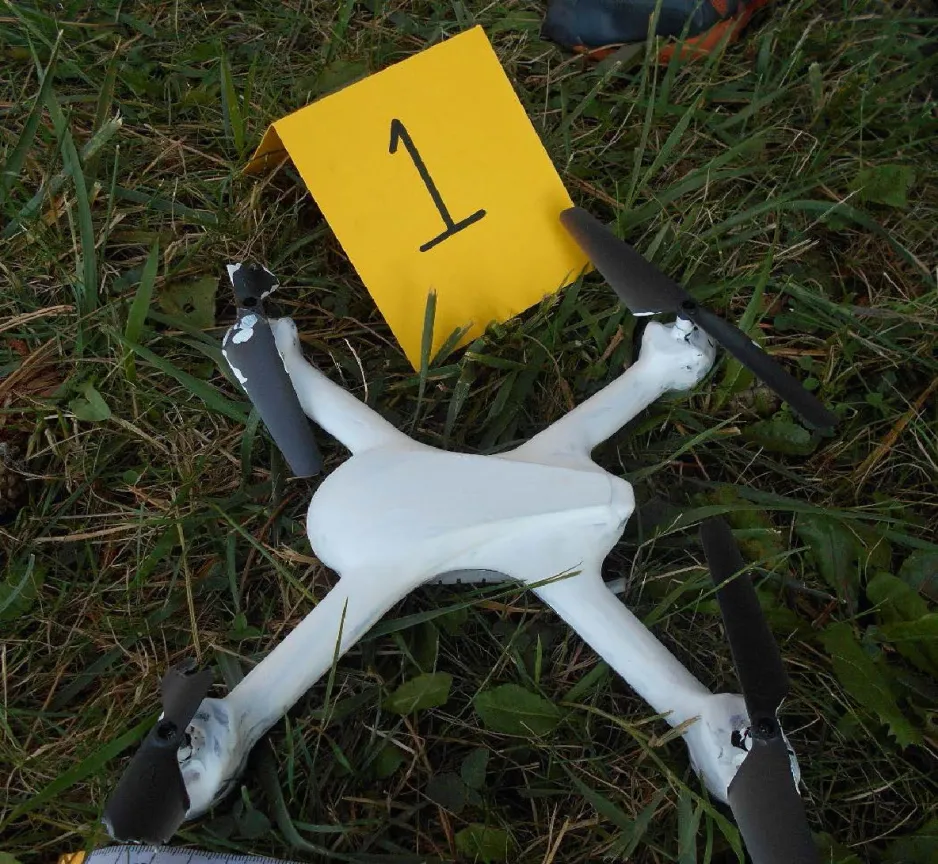 Un drone blanc aux hélices brisées repose sur la pelouse, à côté d’une balise d’indice.