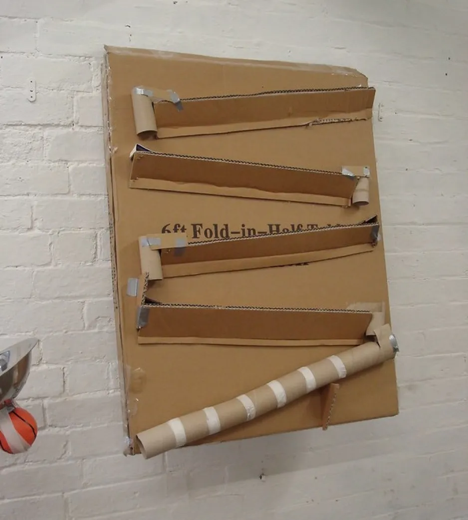Several chutes and paper towel tubes are attached to a large, flat piece of cardboard mounted to a wall. The tubes and chutes are connected to make a continuous pathway for a marble. 