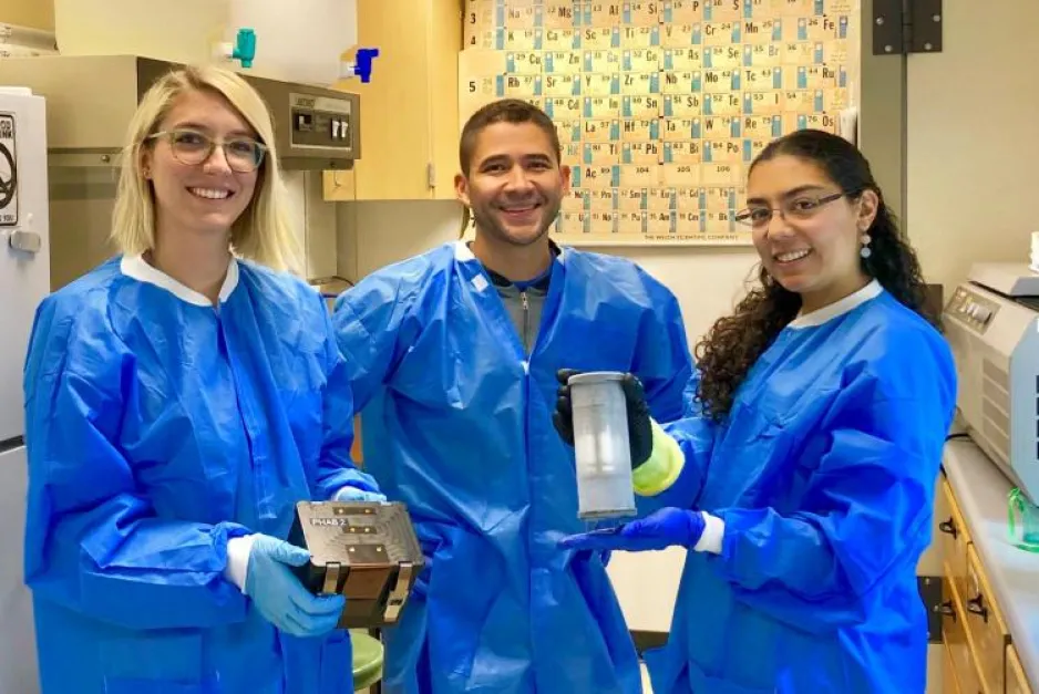 Three researchers wearing blue protective aprons hold equipment that houses bacteria and fungal samples that will be sent to the ISS.