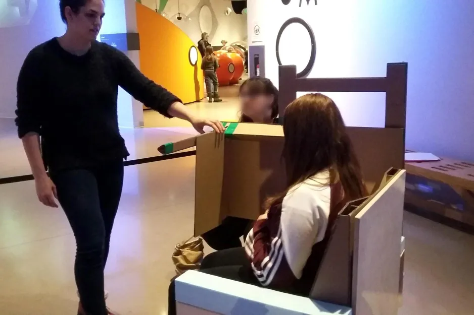 A young woman is seated in a chair inside the museum; another woman stands in front of her.