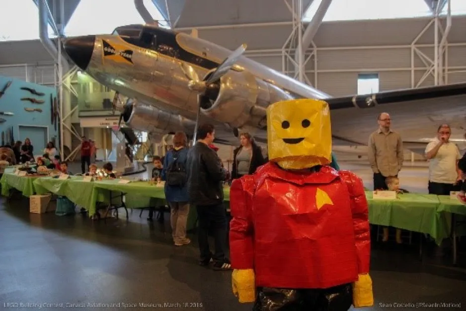 Une personne déguisée en personnage Lego souriant au Musée; on aperçoit un avion à l’arrière-plan.