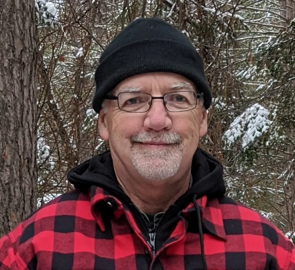 A bearded man with glasses, a black hat, and a red-and-black checkered jacket stands in front of a tree.