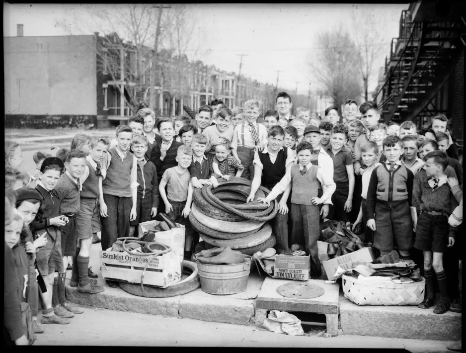 A large group of children, mostly young boys, and a man, are gathered in a semi-circle on a suburban sidewalk, surrounding a pile of car and bicycle tires, and rubber boots which they had collected.