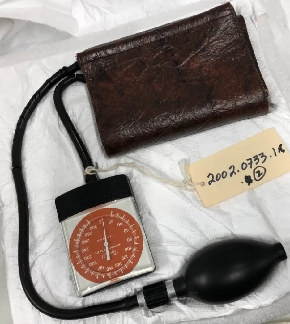 A vintage blood pressure cuff, with an artifact tag attached, sits on a table. 