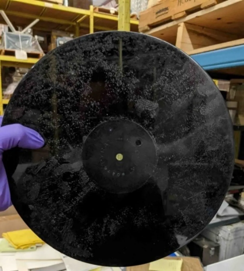 A gloved hand holds up a black disk; greyish-white spots appear on the surface indicating plasticizer that is migrating to the surface.