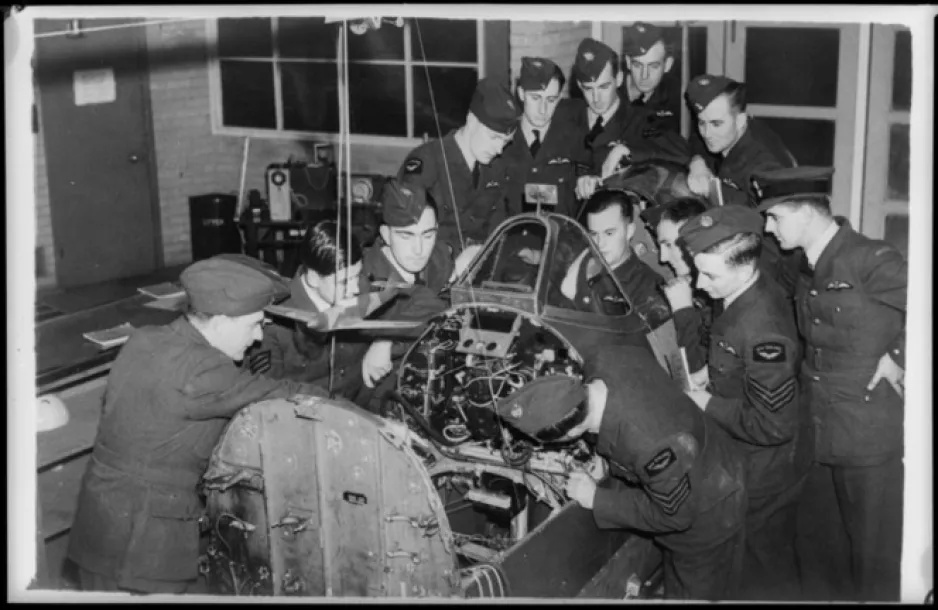 A group of men in air force uniforms surround the detached cockpit section of a Spitfire, looking at how its controls operate.