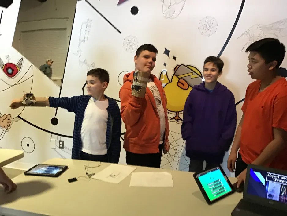 Four boys show off their inventions to museum guest. They have invented wristbands to help people move through airports. Two boys model their prototypes, which are made from cardboard and circuitry. 