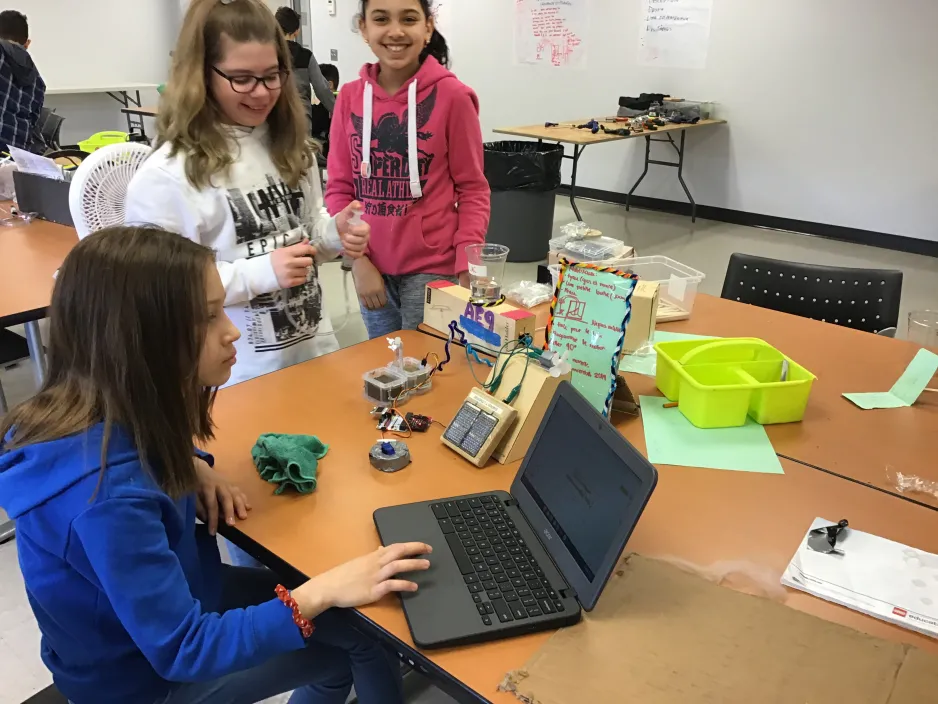 Trois filles programment les circuits pour leur invention. Une variété d'appareils informatiques et de circuits est disposée devant elles. 