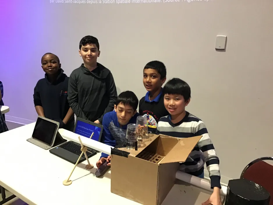 5 garçons montrent leur invention aux visiteurs du musée. Devant eux se trouvent un ordinateur et une boîte en carton avec plusieurs pièces jointes et des fils qui sortent