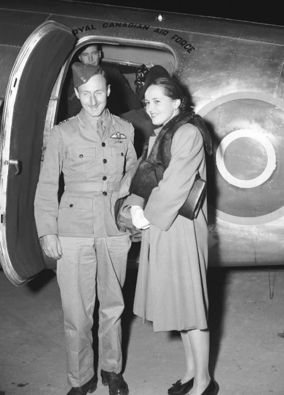 Une image en noir et blanc présente un jeune homme en uniforme militaire, debout près d’une jeune femme portant des vêtements élégants et des talons hauts. Sur l’avion derrière eux on peut lire « Royal Canadian Air Force ».