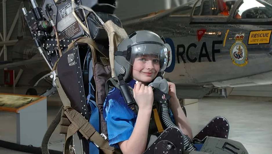 A young boy sitting in a pilot’s seat which has been removed from the aircraft and is sitting on the floor of the museum