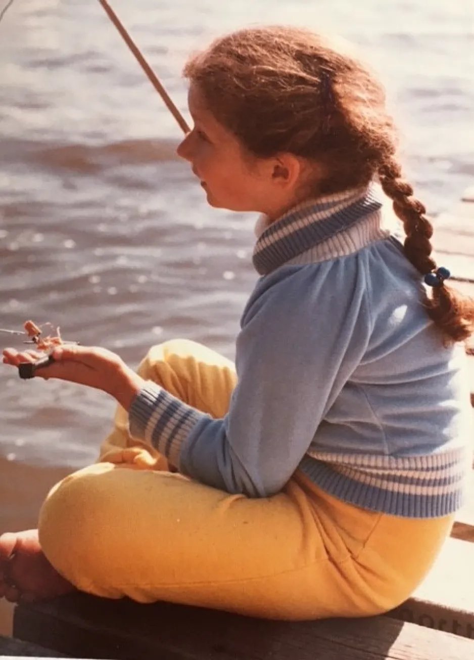 A little girl wearing yellow pants and a blue shirt sits on a dock; the sun sparkles on the lake’s surface next to her. 