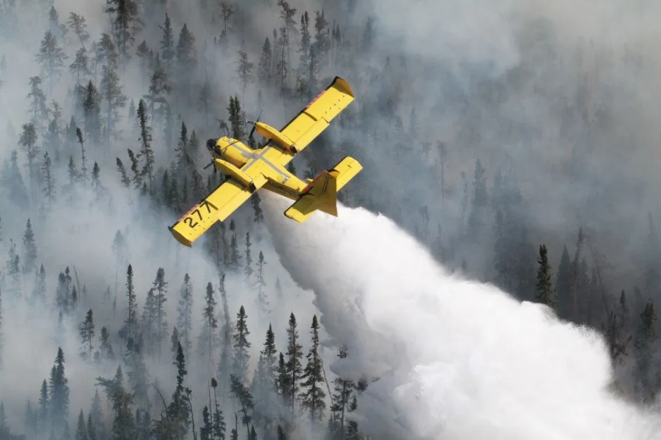 An aerial view of a bright yellow aircraft, a CL-415, flying over the treeline of a forest. The aircraft is spraying water below, while huge clouds of smoke billow over the trees.