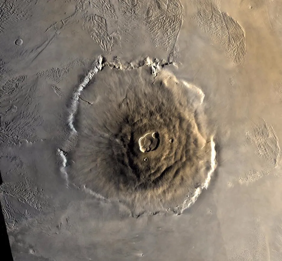 A volcano in the form of a brown wispy-textured mound is centred on a beige, relatively flat lying background. At the centre of the mound are a series of overlapping flat-bottomed craters, or calderas. There are also two smaller impact craters on the flank of the volcano.  At the base of the volcano is an irregularly-shaped terrace ledge.  