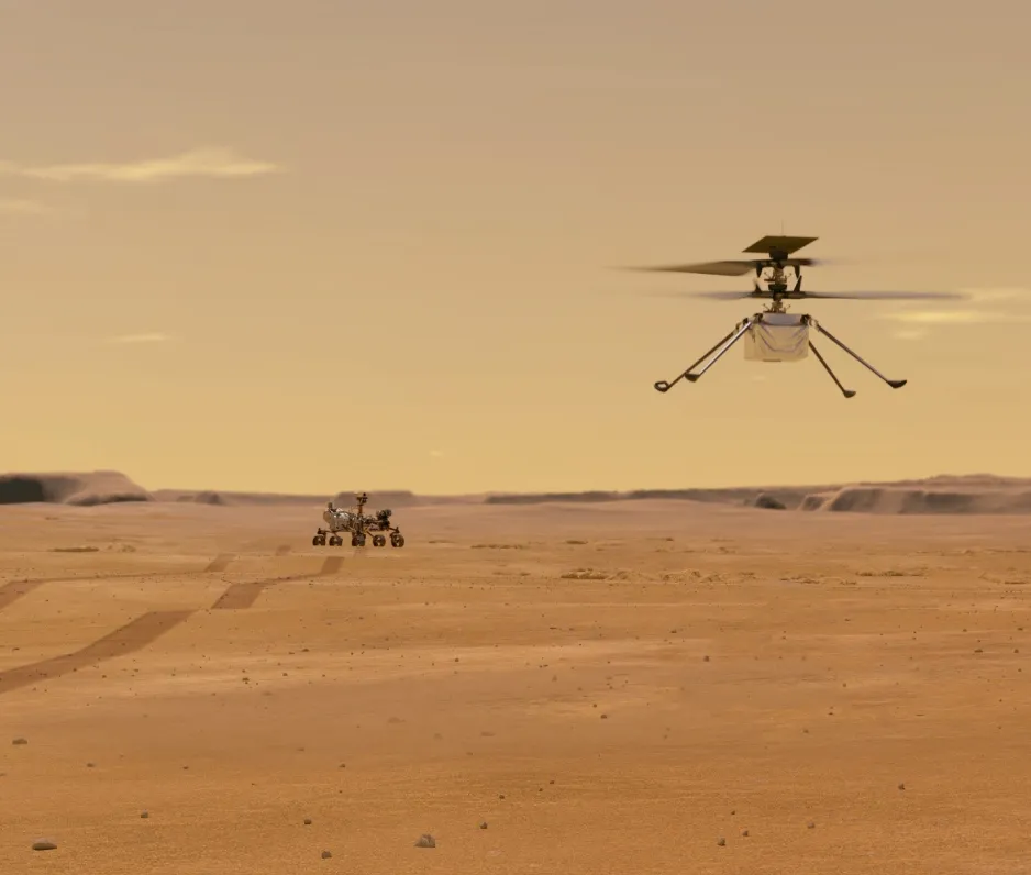 An orange-brown sandy landscape with the Mars Perseverance rover in the background, and rover tracks leading off to the left of the image. The Ingenuity helicopter, a small double-rotor craft with four legs, is flying in the foreground a few metres above the surface.