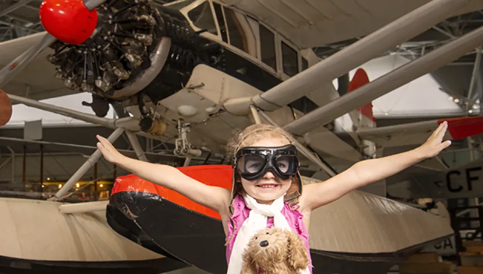 Une jeune fille portant des lunettes d'aviateur, un foulard blanc et un casque d'aviateur brun est assise devant un hydravion dans le musée. Elle a un chien en peluche marron sur ses genoux et ses bras sont tendus comme si elle volait. 
