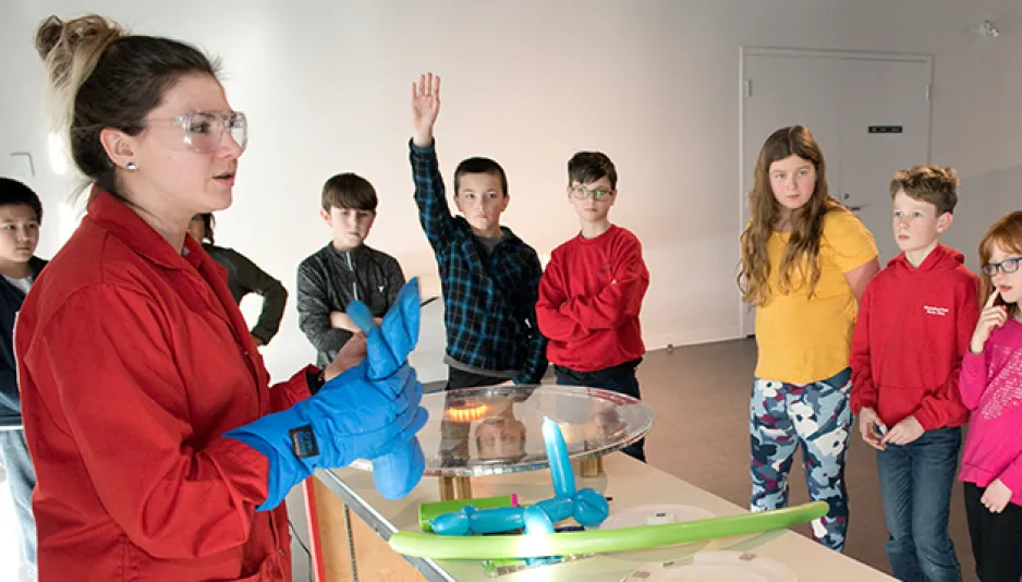 Une personne portant une blouse rouge, des lunettes de protection et des gants bleus se tient devant une table sur laquelle se trouvent divers objets. Un grand groupe d'enfants se tient devant la table et regarde, et deux des enfants ont les mains levées en l'air.