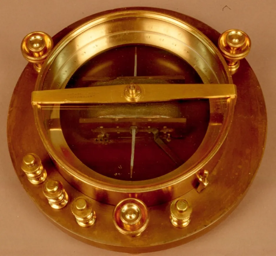 Dispositif en forme d’anneau, de couleur laiton, monté sur une base ronde en bois d’un diamètre légèrement supérieur. Le dispositif comporte une façade en verre et sept petits poteaux en laiton sur son pourtour.