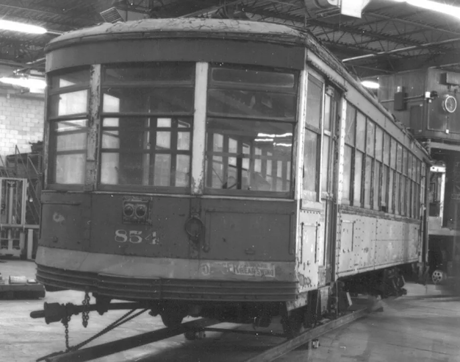 : Photo en noir et blanc d'un vieux tramway portant le numéro 854 à l'avant. Le tramway a des fenêtres sur tous les côtés et repose sur des rails de tramway temporaires dans un entrepôt.