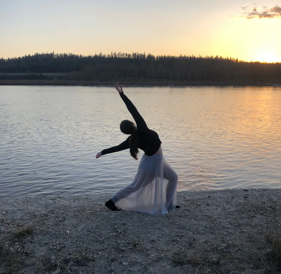 Une femme portant un haut foncé et une jupe fluide translucide prend une pose de danse près d’une rivière. Le soleil est bas sur l’horizon. 