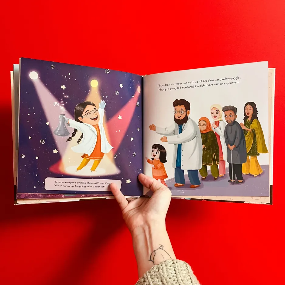 A hand holding an open children's book, on the inside left of the book it depicts an illustration of a young girl in a lab coat excited with her hands in the air holding a beaker, on the right it shows a family watching and smiling at the young girl.