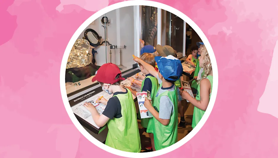 A group of children wearing green pinnys stand in front of a museum exhibition showcase which contains examples of different products that are made with glass.  Some children point at the showcase while others write down information on the activity sheets they are holding.