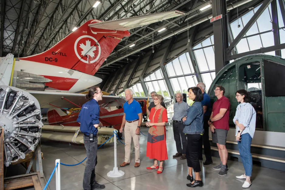 A group of people smile while listening to a museum guide speak about the museum's aircraft collection. The tail of an Air Canada aircraft is visible in the background
