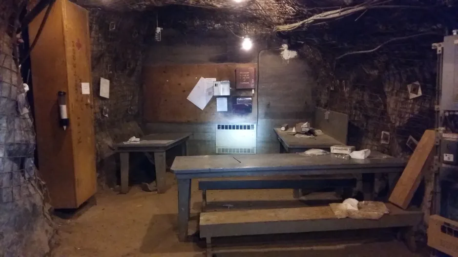 An underground refuge chamber is equipped with a table and benches. The space is dimly lit by several small light sources. 