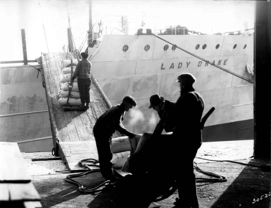 A black and white photograph showing a man pushing a cart of flour sacks up a plank leading to the deck of a large ship. The ship has the words “Lady Drake” written on the side. In the foreground, the silhouettes of three men prepare more flour sacks for loading onto the vessel.