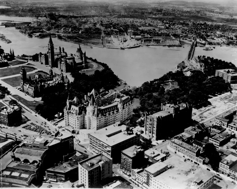 A black and white photograph taken from the air, showing a river flanked by buildings. The tall, stone Parliament Buildings are in the top left corner of the photo, with a canal to their right, and another large, stone building with turrets and spires to the right of the canal.  The image also contains a number of tall, rectangular structures ranging from 4 to 10 storeys tall.