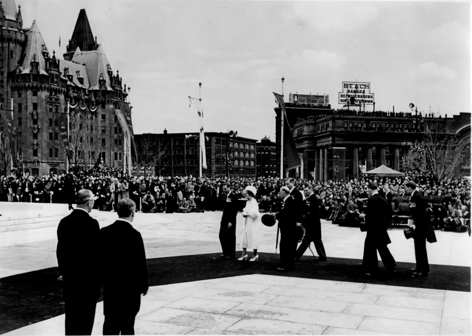 Photo noir et blanc d’un homme et d’une femme en tenues élégantes, marchant devant plusieurs hommes en costume, sur un tapis de cérémonie installé sur le pavé. Une grande foule se trouve à l’arrière-plan.