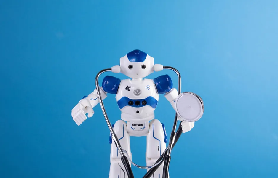 A humanoid toy robot wears a stethoscope, against a light blue background. 