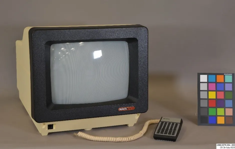 A cathode ray tube computer terminal and small keyboard sitting on a grey background. The computer has yellow plastic housing and black plastic frame, and the keyboard is grey with black keys and a yellowed spiral plastic cord. On the right side of the picture, there is a conservation photography colour correction card.
