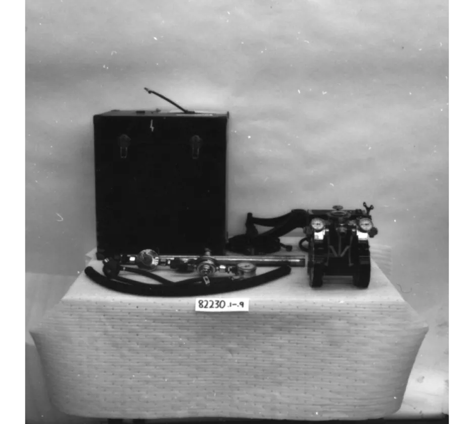 A black-and-white image depicts a large rectangular tank, a mask, hose, and some metal pieces lying on a table.