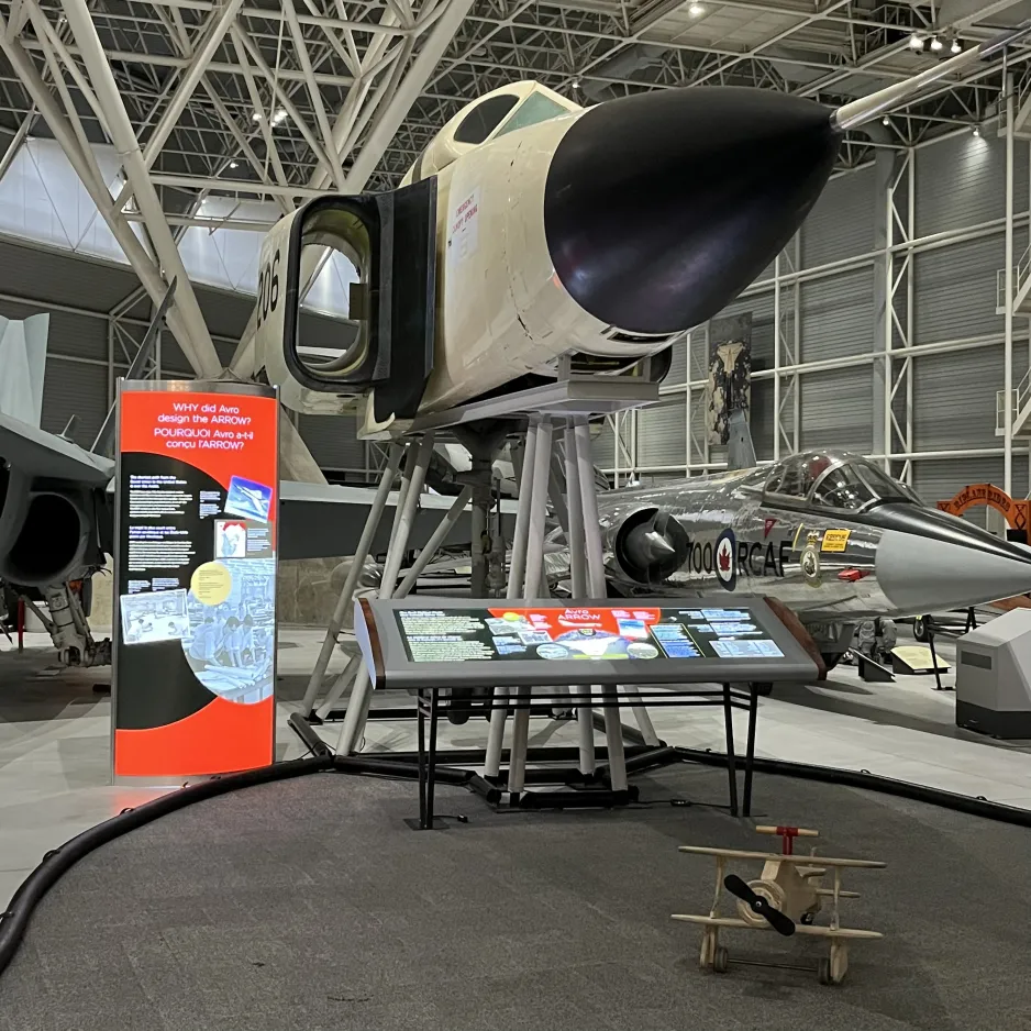 View facing the nose section of the Avro Arrow. A new backlit console panel sits below the nose: black with grey and vivid red highlights. A tall upright panel sits to the left of the nose, also backlit, with a similar black and red design. A small wooden wheeled biplane, the type that a small child would ride upon, sits in the foreground.