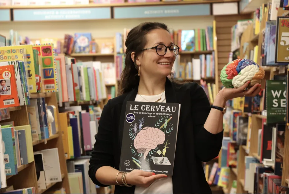 A person with shoulder length hair is standing in a store surrounded by book shelves and is holding a book called "Le cerveau" in one hand and a brain model in the other as they stare at it.