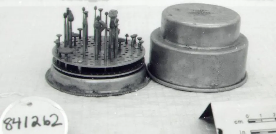A round, metal tray has holes that hold an array of metal drill bits. A lid for the collection sits next to the drill bits on a light-coloured backdrop.