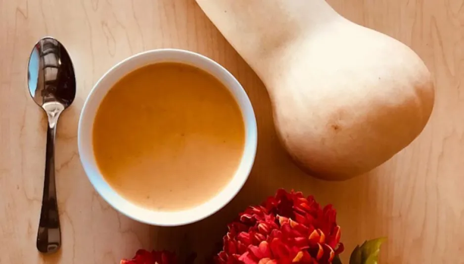 A bowl of soup and a spoon sit on a wooden surface. A butternut squash and a flower are also visible.