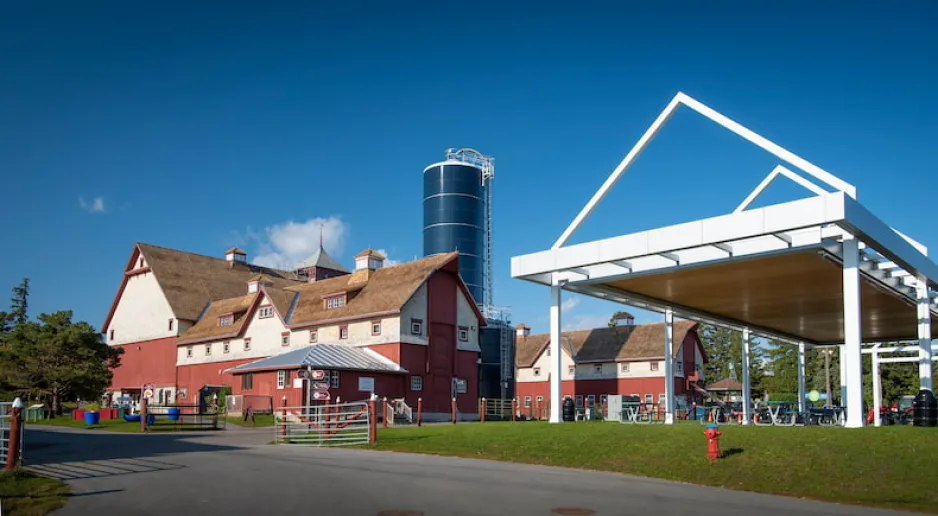 A wide shot of two large, red-and-white agricultural buildings and a navy blue silo. Green grass and a covered, outdoor picnic area are visible in the foreground, and a blue sky in the background.