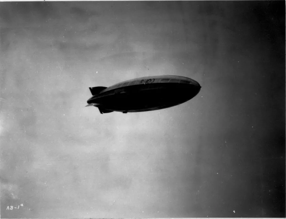 A black-and-white photograph taken from the ground looking up at a large, oval-shaped blimp-like airship flying overhead.