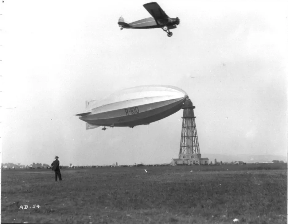 A black-and-white photograph showing a small, propeller-driven airplane flying over a large, oval-shaped blimp-like airship, tied to the top of a tall tower that looks like a lighthouse. A man stands in the foreground looking on.