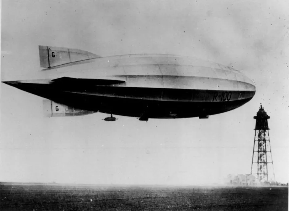 A black-and-white photograph of a massive blimp-like airship, tied to the top of a tall tower that looks like a lighthouse.