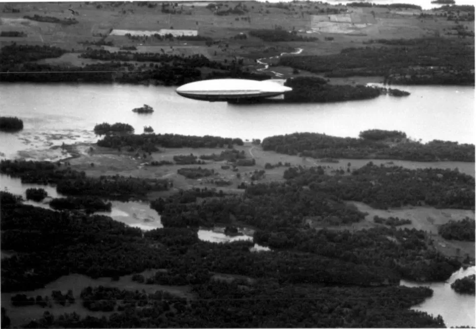 A black and white photograph of a massive blimp-like airship flying over a river and islands below. This photograph was taken from an aircraft flying near the R-100.
