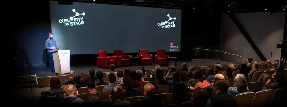 Jeff Yates stands behind a podium, on a stage, with a four armchairs and a screen behind him. He stands at the front of an auditorium filled with people.  