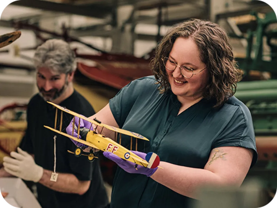 Une personne aux cheveux mi-longs et aux lunettes examine un modèle réduit d'avion. Un collègue aux cheveux gris et à la barbe regarde à l'arrière-plan.